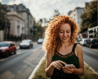 Rothaarige Frau steht mitten auf der Straße und blickt in ihr Handy und lächelt dabei