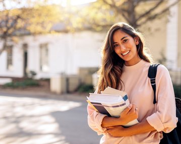 Studentin mit langen braunen Haaren und rosafarbenem Pullover steht mit Büchern in der Hand vor dem Gebäude